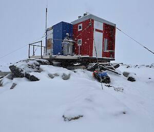 A small cabin atop a wooden platform over some snow-covered rocks. The cabin's walls are painted bright red, with a blue-painted cold porch jutting out from the front. The hut is anchored by guywires to its platform and to unseen fixtures beneath the snow