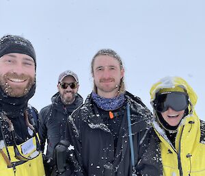 Four people in polar weatherproof clothing stand together, smiling for the camera. Their clothes and hair are lightly dusted with snow