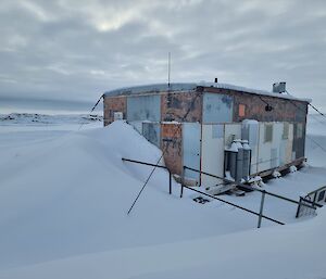 A weatherbeaten hut with a signboard reading "Wilkes Hilton" stands in a small hollow in the snow. A big build-up of snow at one end of the hut blocks the entrance