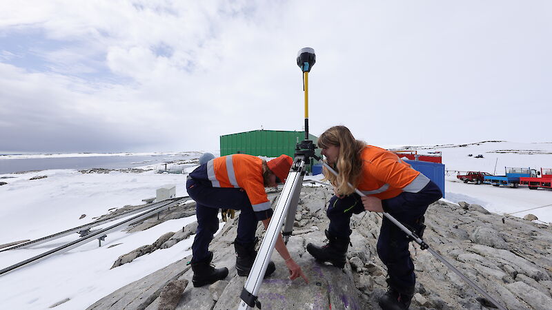 Two scientists stand on a rocky outcrop doing survey work. The Antarctic background is snowy, with the ocean in view