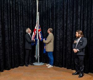 Two men lowering the Australian and New Zealand flags on a short flagpole set up in an indoors location. A third man with military medals pinned to his jacket stands nearby