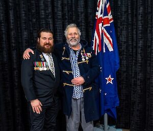 Two men with military medals on their jackets, standing next to an indoor flagpole with their arms around each other's shoulders and smiling