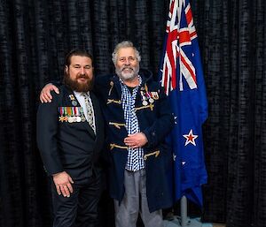 Two men with military medals on their jackets, standing next to an indoor flagpole with their arms around each other's shoulders and smiling