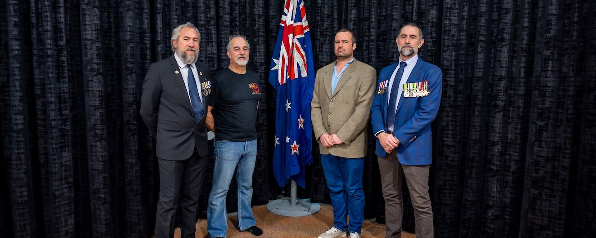 Four men stand in front of a indoor flagpole with the Australian and New Zealand flags at half-mast. Two of the men wear military medals on their jackets