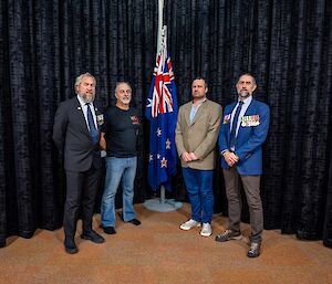 Four men stand in front of a indoor flagpole with the Australian and New Zealand flags at half-mast. Two of the men wear military medals on their jackets