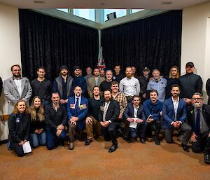 A group of about 30 people pose for a photo, standing and kneeling in rows before an indoors flagpole bearing the Australian and New Zealand flags