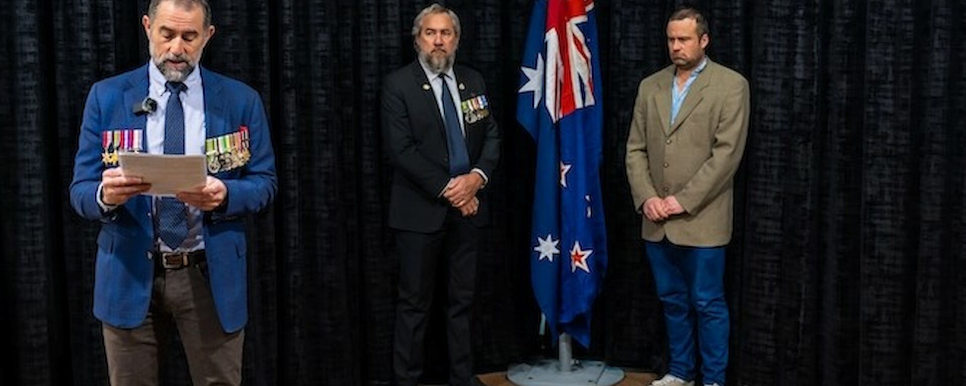 Two men stand on either side of an indoor flagpole bearing the Australian and New Zealand flags. A man with military medals on his jacket stands in front of them, reading aloud from a programme