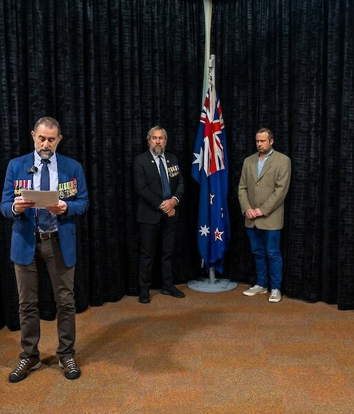 Two men stand on either side of an indoor flagpole bearing the Australian and New Zealand flags. A man with military medals on his jacket stands in front of them, reading aloud from a programme