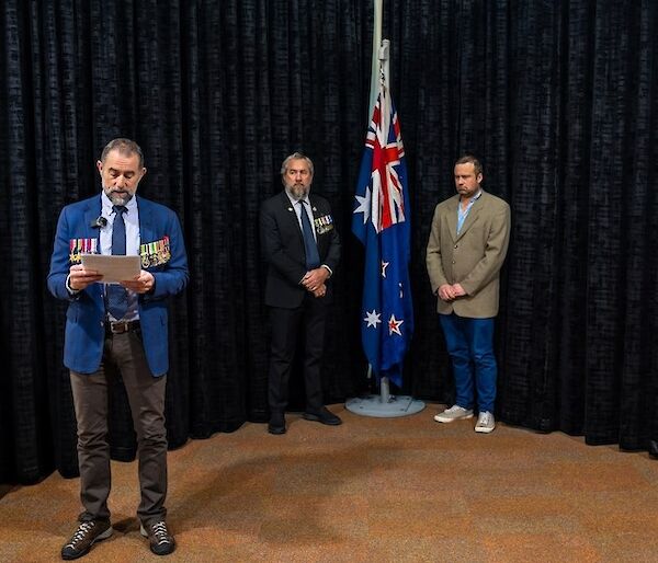 Two men stand on either side of an indoor flagpole bearing the Australian and New Zealand flags. A man with military medals on his jacket stands in front of them, reading aloud from a programme