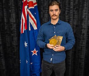 A solemn young man stands in front of an indoor flagpole, holding a medallion and an old, sepia-toned photograph of a man in ANZAC uniform