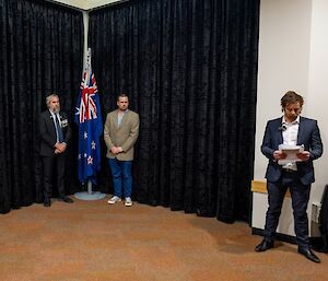 Two men stand on either side of an indoor flagpole with flags lowered. Another man standing to the side reads aloud from a programme