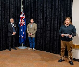 Two men stand on either side of an indoor flagpole with flags lowered. Another man standing to the side reads aloud from a programme