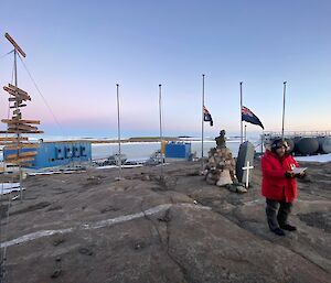 Man stands in foreground reading from pamphlet, behind are Australian and NZ flags at half mast and a commemorative cross, in distance Horseshoe Harbour