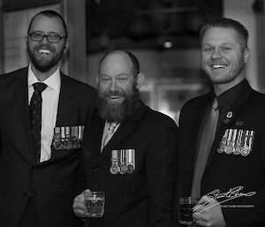 Black and white photo, three men in suits and ties wearing service medals smiling to camera
