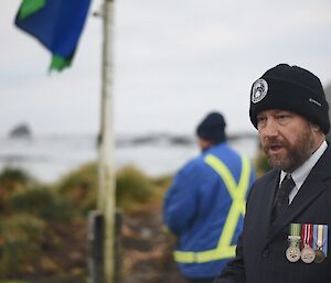 Man in foreground in suit and tie wearing AAD beanie and service medals, in background man standing at flag pole with flag at half mast