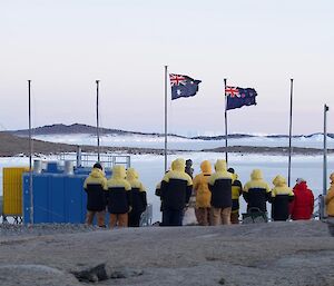 Group of expeditioners gathered around the mawson flag poles with ice covered harbour in distance