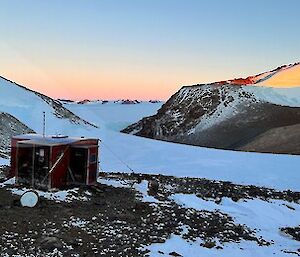 Small square field hut in foreground, overlooks snow covered valley between two rocky peaks, in the distance across the ice plateau is mountain range at sunrise