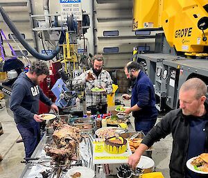 Men gather around workshop bench with is covered with food - lamb spit, salads, and birthday cake.