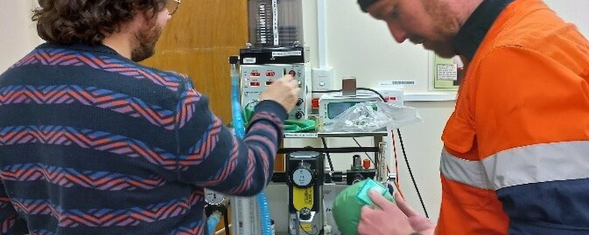 Two people in a medical facility are setting up the ventilator by checking the tubes, wires and dials