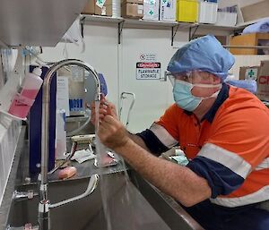 A person undertakes hand scrubbing pre surgery over a stainless steel sink.  He wears a head scarf and face mask