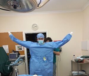 A man stands arms out while he dons a blue sterile gown in a medical facility, wearing a head scarf and face mask
