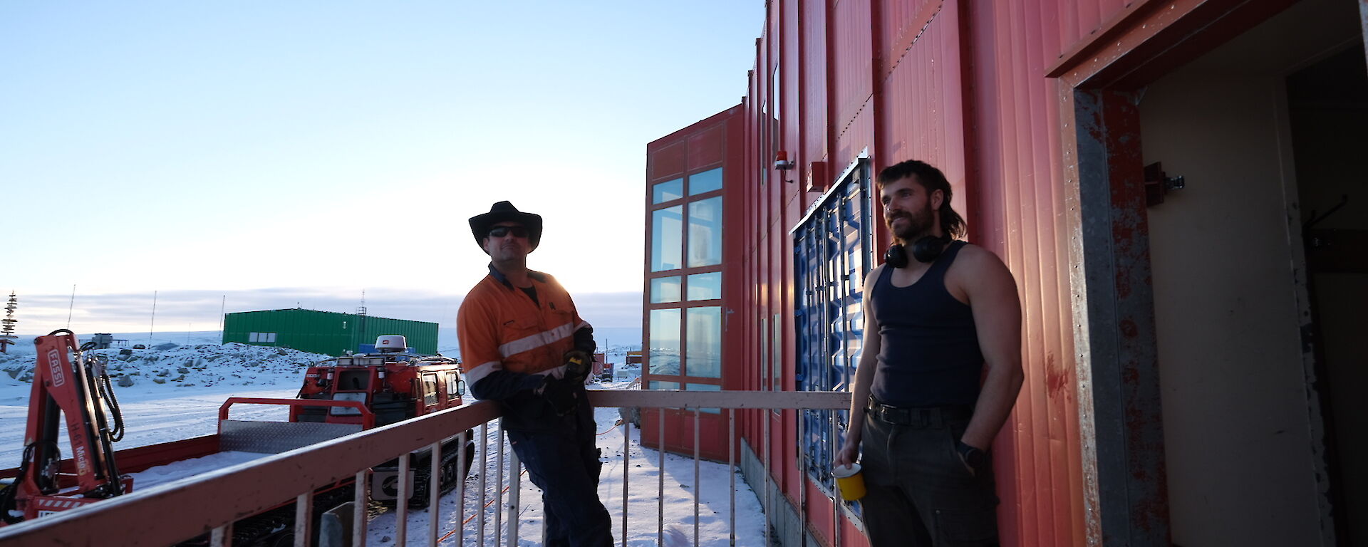 Two men standing on a metal grille landing attached to a tall building with a red-painted metal exterior. A tracked vehicle with a flatbed in tow is parked on the snowy ground behind them. The sky is soft blue in the early morning light