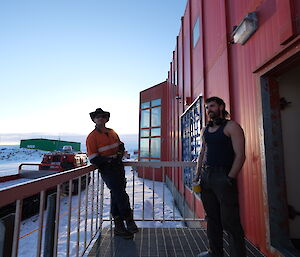 Two men standing on a metal grille landing attached to a tall building with a red-painted metal exterior. A tracked vehicle with a flatbed in tow is parked on the snowy ground behind them. The sky is soft blue in the early morning light