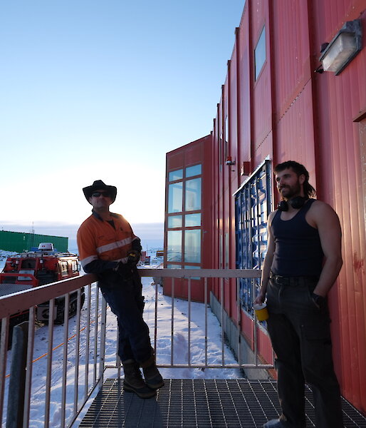 Two men standing on a metal grille landing attached to a tall building with a red-painted metal exterior. A tracked vehicle with a flatbed in tow is parked on the snowy ground behind them. The sky is soft blue in the early morning light