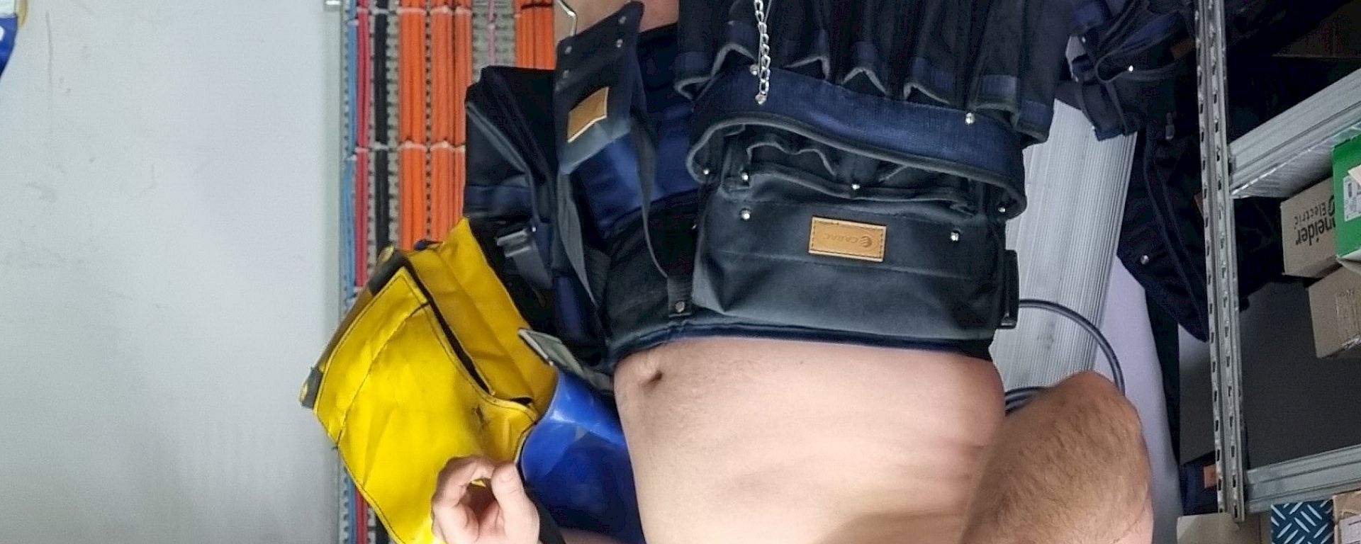 A man in a workshop wearing nothing but work boots, a wide and covering tool belt and a sheriff hat, pointing his fingers like a gun to strike a comical pose