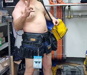 A man in a workshop wearing nothing but work boots, a wide and covering tool belt and a sheriff hat, pointing his fingers like a gun to strike a comical pose