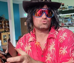 A man in a red Hawaiian shirt patterned with yellow palm trees, an unruly black-haired wig, reflective sunglasses and a sheriff hat sitting at a counter, pouting for the camera