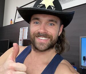 A man in a blue singlet and sheriff hat, smiling and giving a thumbs-up to the camera