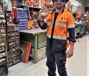 A man in high visibility work clothes standing in a workshop, wearing a sheriff's hat and sunglasses