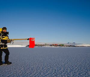 Man stands on sea-ice holding canes with red flags on end. In the distance is Mawson station