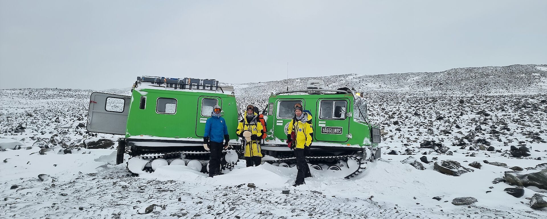 3 men standing in front of a Hagglunds surrounded by rocks and snow