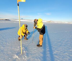 Two men preparing to measure thickness of sea-ice, one holds large drill with bit approx 1m long and other is kneeling beside preparing to drop measuring tape through the hole