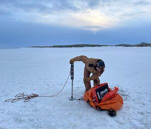 Man holding a large pole with camera attached which is inserted through a hole in the ice is looking at a computer screen resting on large orange duffle bag sitting on ice beside the hole.