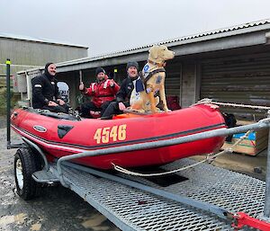 A red boat is on the back of a trailer with a replica of a dog on the front.