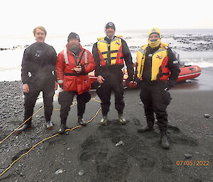 Four people in boating attire stand on the grey foreshore