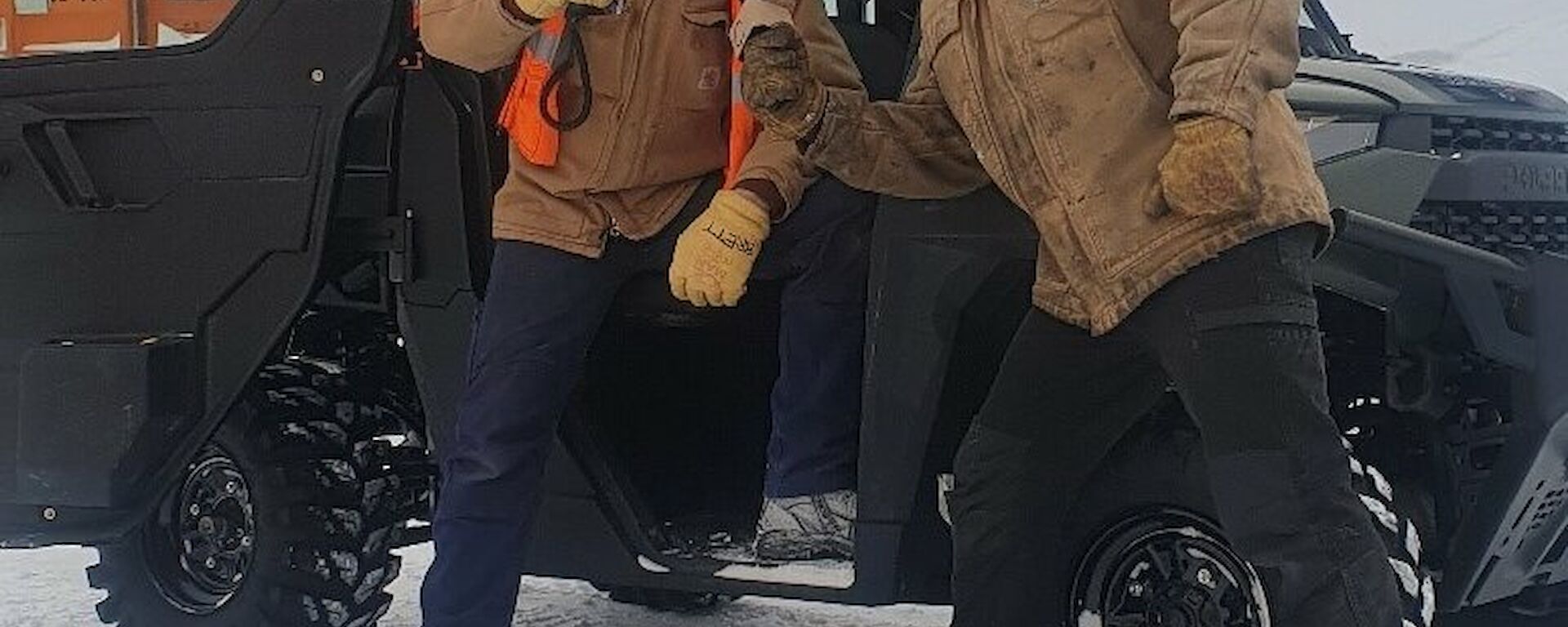 A man in a Carhartt jacket and thick gloves is climbing out of a Polaris vehicle parked on snowy ground, while another man holds a mock microphone towards him, pretending to conduct a TV interview
