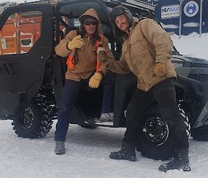 A man in a Carhartt jacket and thick gloves is climbing out of a Polaris vehicle parked on snowy ground, while another man holds a mock microphone towards him, pretending to conduct a TV interview