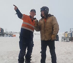 On open, snowy ground, a man in high-vis work clothing points his finger at an unspecified object in the distance. He appears to be showing something to another man standing beside him, who is carrying a mock microphone, pretending to be a TV journalist