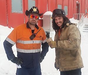 In a snowy location in front of a red-painted building, a man in high-vis work clothes, reflective sunglasses and a sheriff hat strikes a business-like pose. Another man stands beside him with a mock microphone, pretending to conduct a TV interview