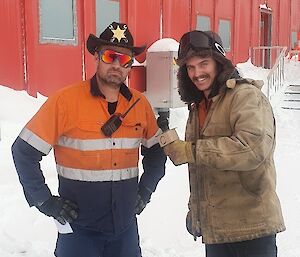 In a snowy location in front of a red-painted building, a man in high-vis work clothes, reflective sunglasses and a sheriff hat strikes a business-like pose. Another man stands beside him with a mock microphone, pretending to conduct a TV interview