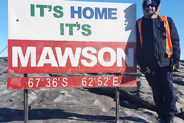 Man stands beside a sign which reads "it's home, it's Mawson" and latitude and longitude of 67deg 36secs S and 62dec 52 secs E.