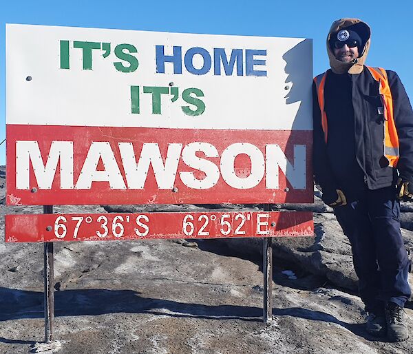 Man stands beside a sign which reads "it's home, it's Mawson" and latitude and longitude of 67deg 36secs S and 62dec 52 secs E.