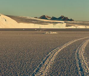 Hagglunds tracks in the sea ice leading back towards ice plateau with mountains in the distance and full moon above in clear blue sky