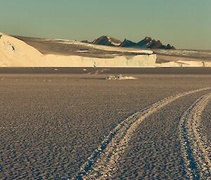 Hagglunds tracks in the sea ice leading back towards ice plateau with mountains in the distance and full moon above in clear blue sky