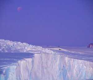 Full moon in partial eclipse over ice plateau as the sun beings to rise giving hues of pink and purple