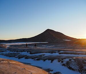 Two people walking across rocky snow covered ground with hill silhouetted behind and sun shining out from behind hill